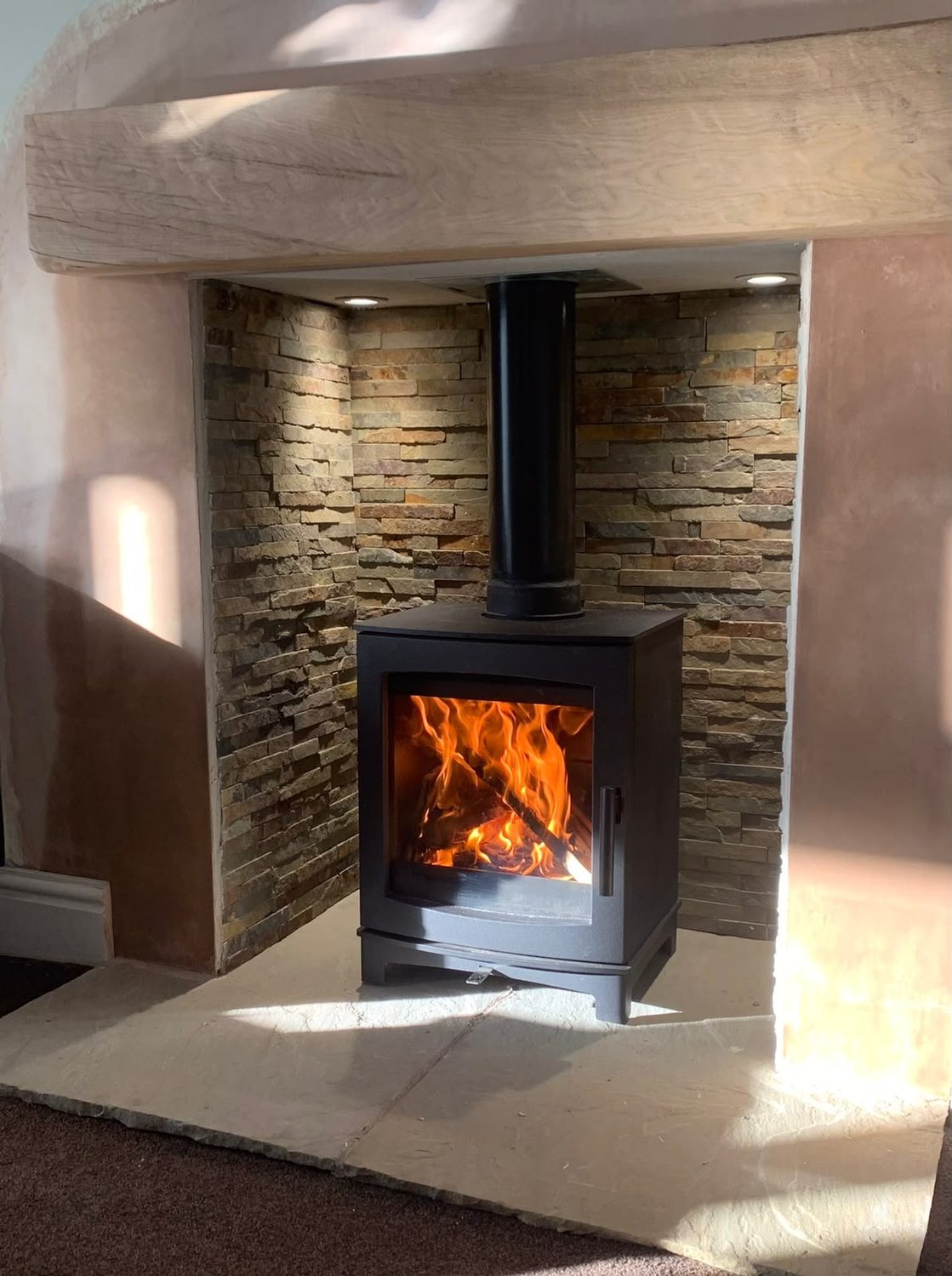 Woodburning stove lit in a split-face stone chamber with an oak beam, fitted by Heavenly Fireplaces in Blackpool