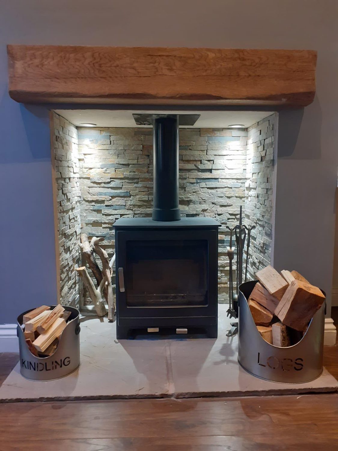 Stove in a downlit split-face stone chamber with log and kindling buckets